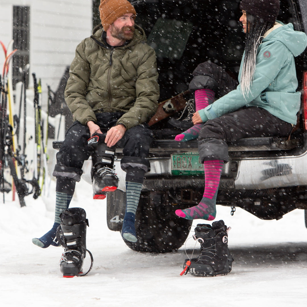 A skier and a snowboarder seated on tailgate comparing socks from darn tough