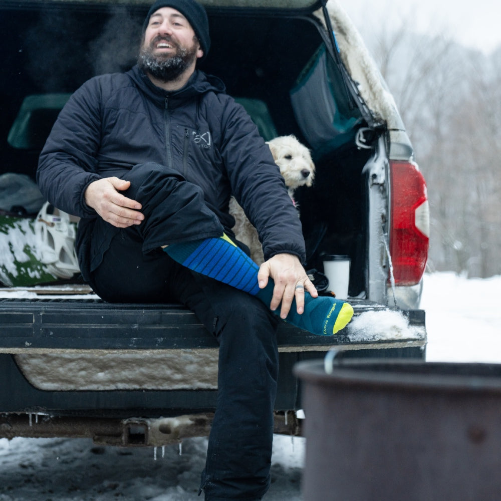 A skier seated on tailgate putting boots on, wearing thick ski socks with cushion