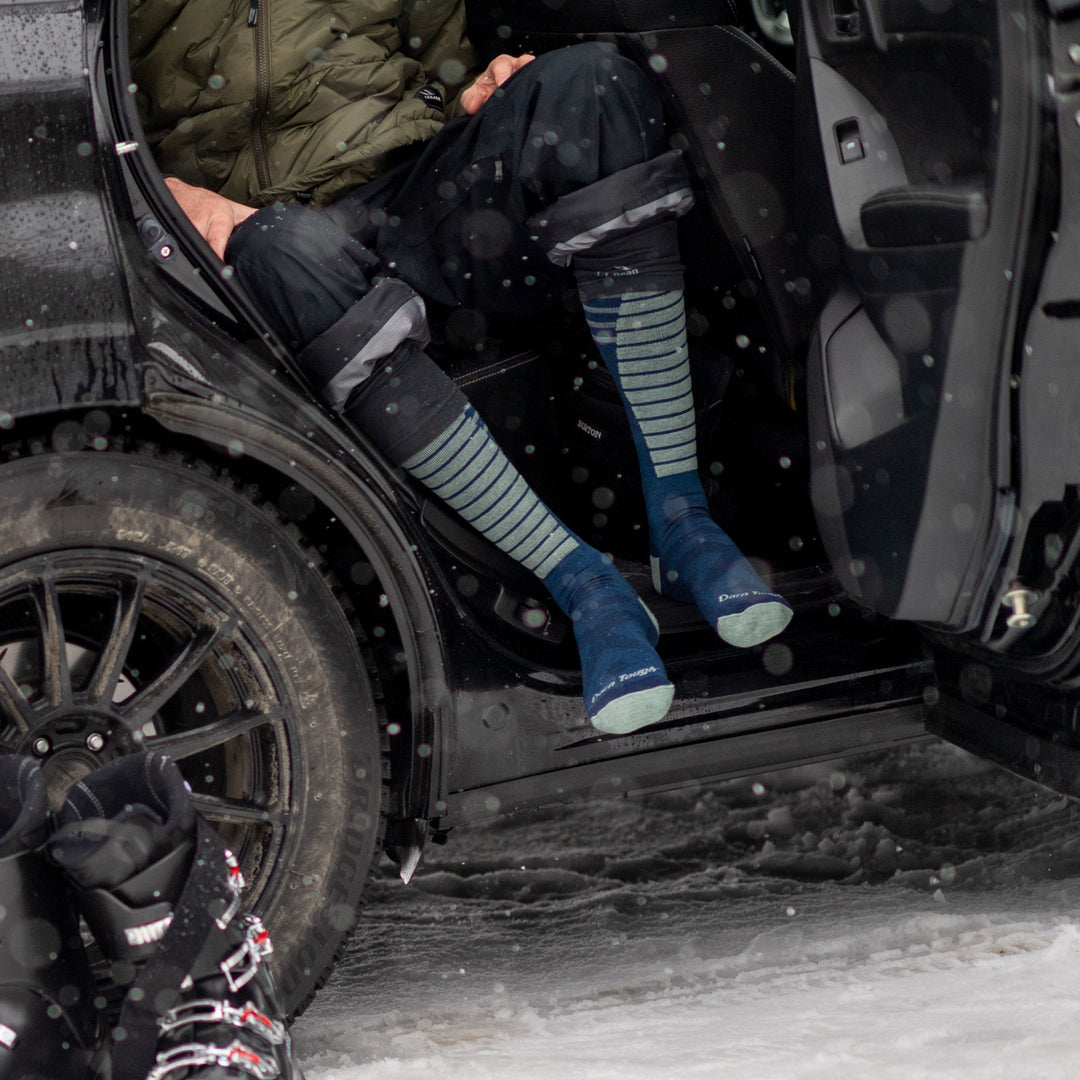 Model sitting in a black car with the door open wearing men's edge over-the-calf snow sock in midnight blue