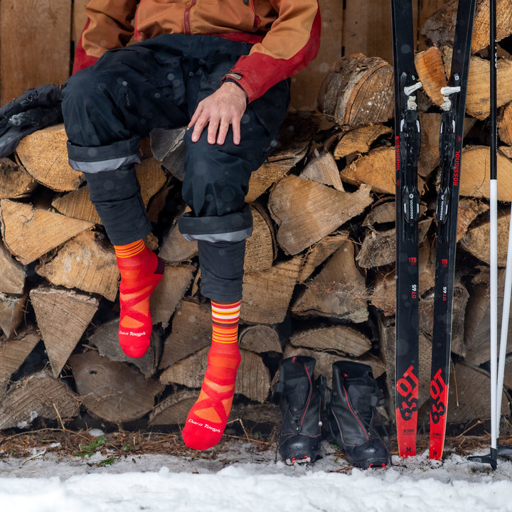 Close up shot of model sitting on log pile wearing men's oslo nordic boot snow sock in rune red with black and red ski