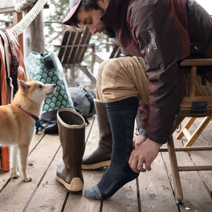 Side shot of model sitting in a wooden chair outside putting on the men's stanley k mid-calf work sock in gravel