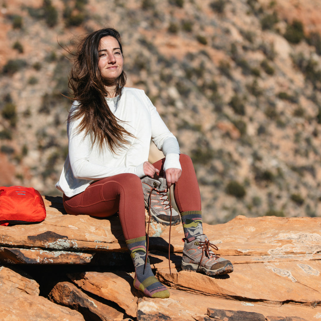 Image of model sitting on red desert rocks wearing 5016 socks in Taupe colorway while wearing one gray hiking boot and holding the other