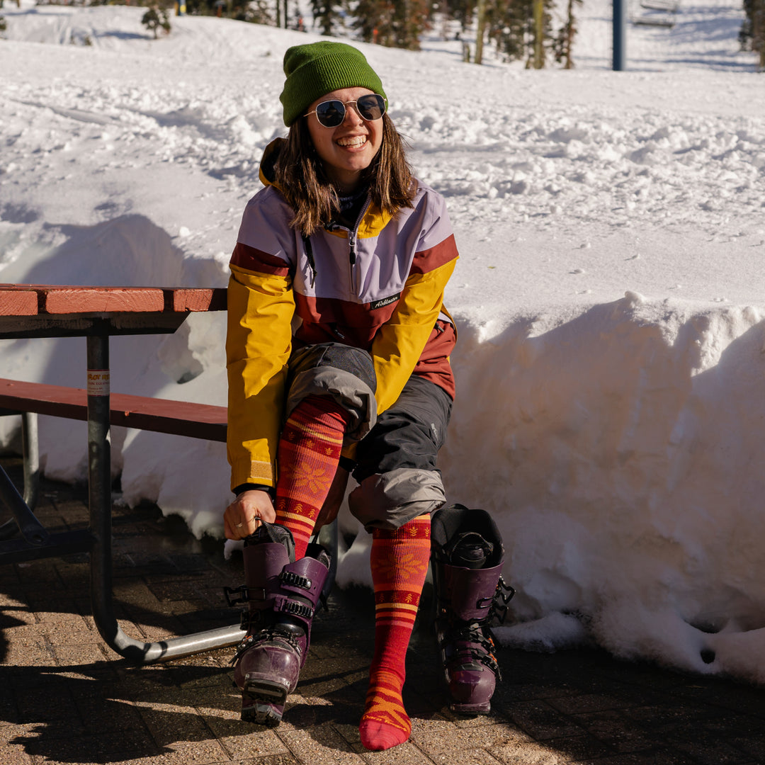 Woman putting ski boots on over her Women's Alpine Over-the-Calf Lightweight Ski and Snowboard Sock in Burgundy
