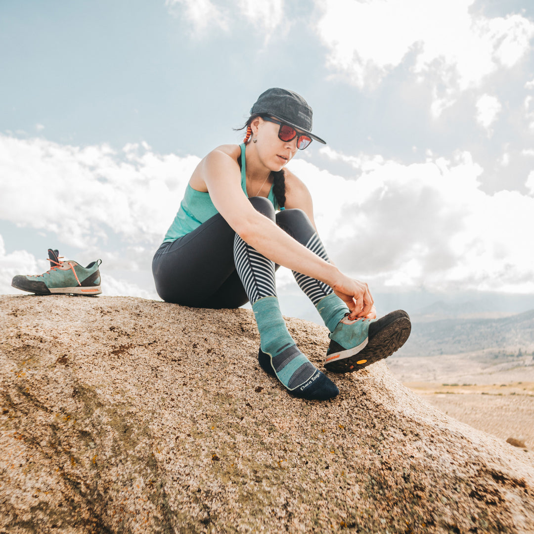 Image of a woman in hiking clothes, sitting on a rock and putting on shoe on while wearing Women's Light Hiker Micro Crew Hiking Socks in Aqua, Lifestyle Image