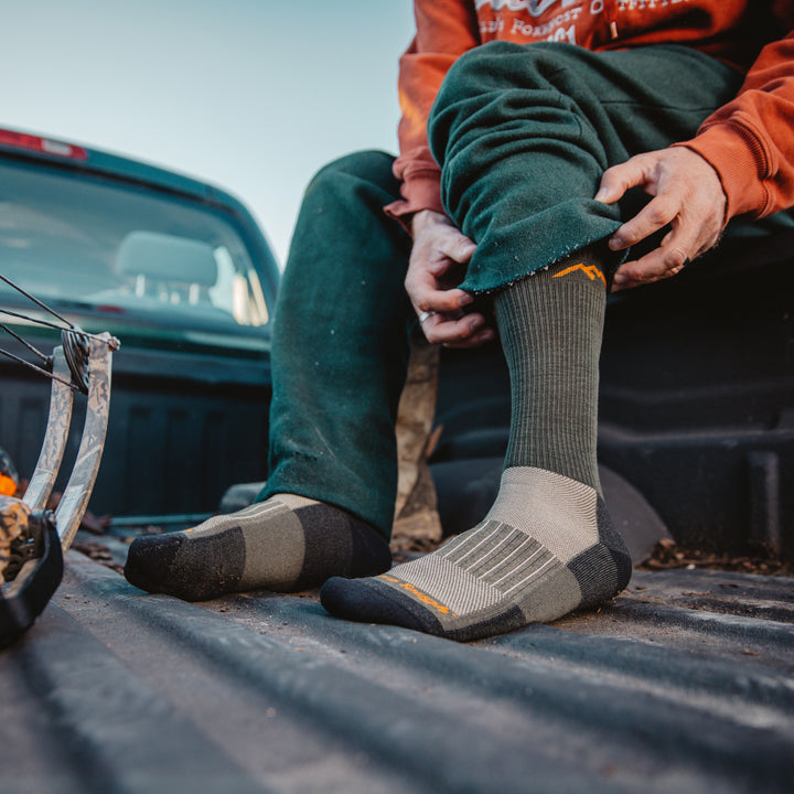 Close up shot of model sitting in a truck bed putting on the men's boot hunting sock n forest green