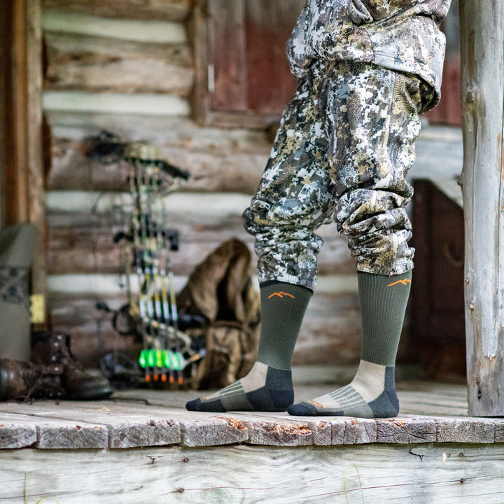 Waist down shot of model standing on wood porch in camouflage wearing the men's boot hunting sock in forest green