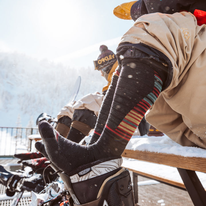 Close up shot of model sitting on a bench outside in the snow wearing the men's backwoods over-the-calf snow sock in charcoal