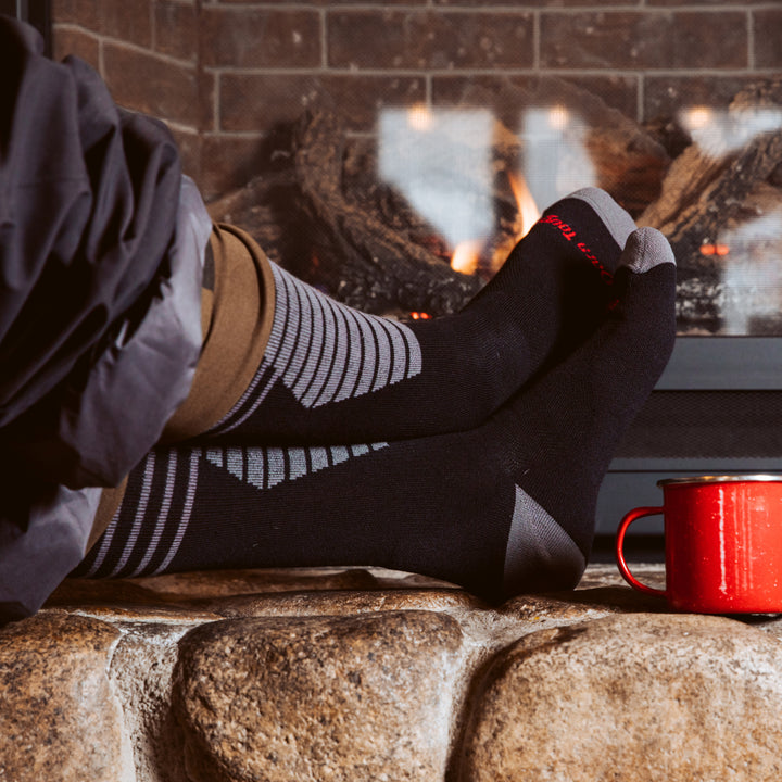 Close up shot of model sitting by a fireplace with their feet up wearing the men's edge oveer-the-calf snow sock in black