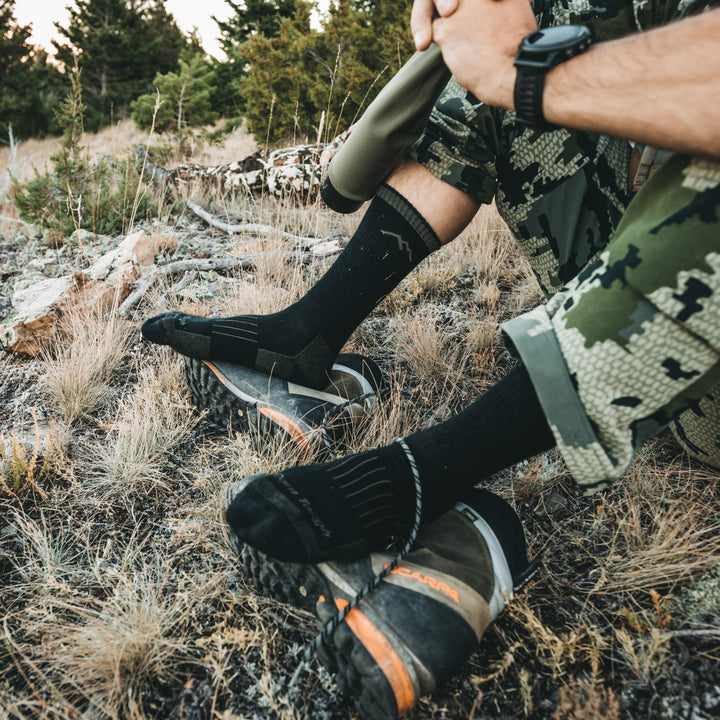 Close up shot of model sitting in the grass in camouflage wearing the unisex boot hunting sock in charcoal