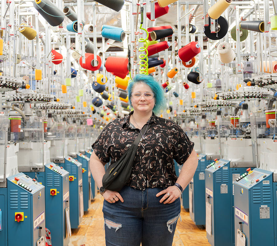 Darn Tough employee Ash standing on the Mill floor, surrounded by knitting machines.