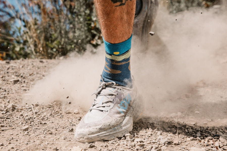 A close-up of a runner's foot mid-stride wearing wool run socks with a western landscape
