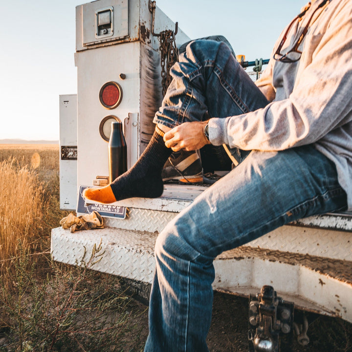 Image of a man sitting on the back of a work truck pulling on his Steely Micro Crew Midweight Work Socks in Graphite, Lifestyle Image