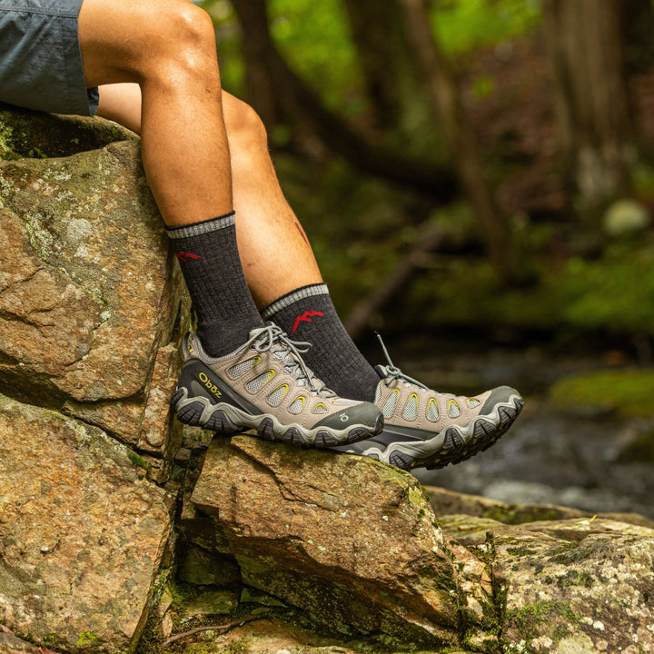 Side shot of model sitting on river rocks wearing the men's micro crew hiking sock in black with brown hiking shoes on