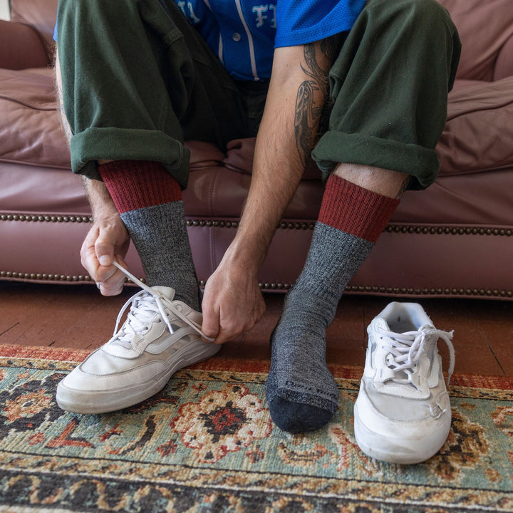Front shot of model sitting on brown couch wearing men's scout boot hiking sock in pepper gray with putting on white sneakers