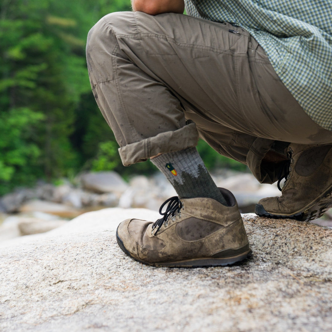 A hiker crouched on a rock wearing the Number 2 Hiking Sock with a rocket outhouse on it