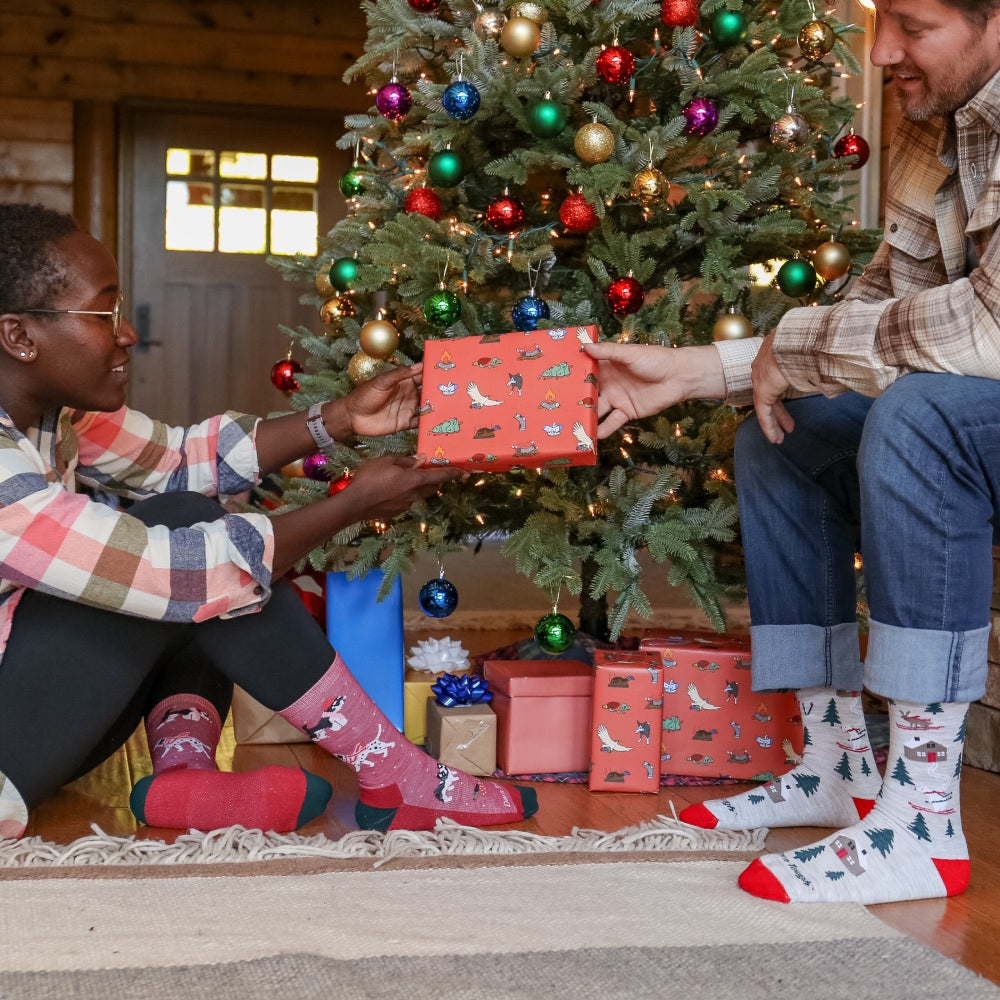 Two people under a Christmas tree, one person passing the other a gift (of Darn Tough socks)