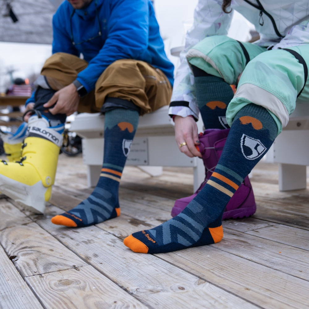 A skier and a snowboarder each wearing over the calf darn tough socks, putting on their boots