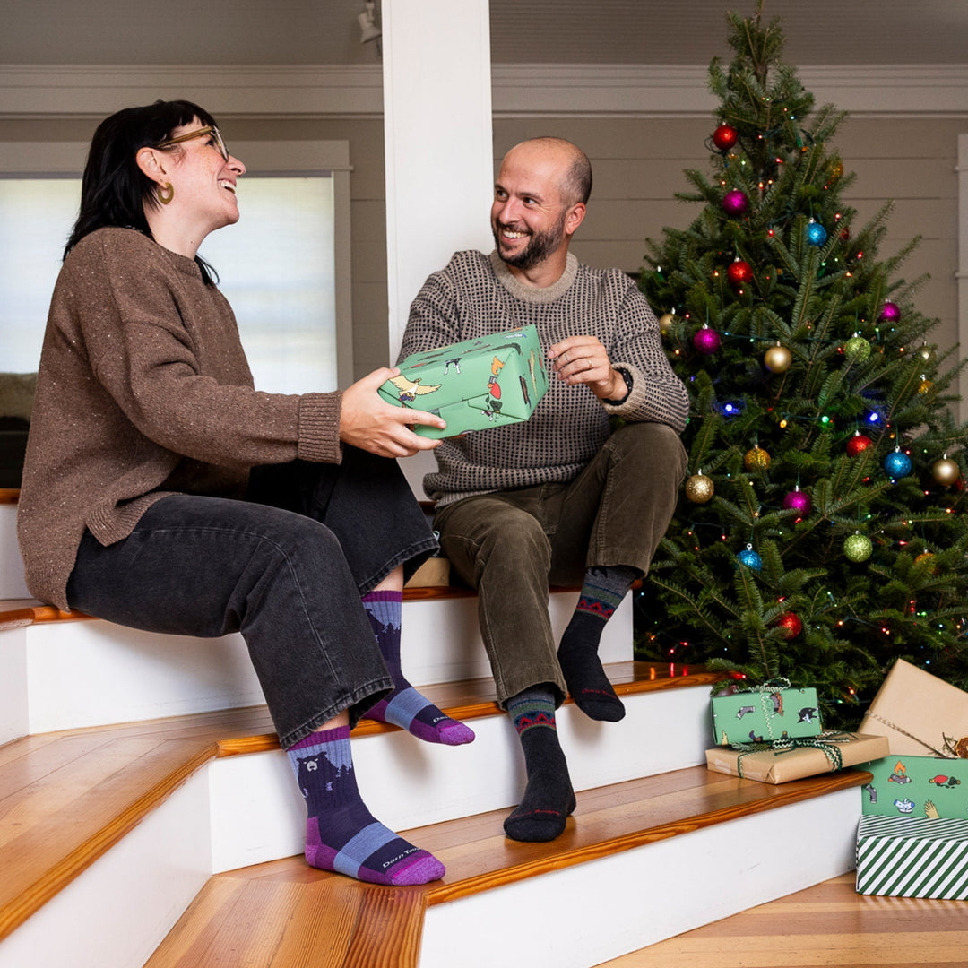 Two people gifting by a Christmas tree, both wearing Darn Tough socks