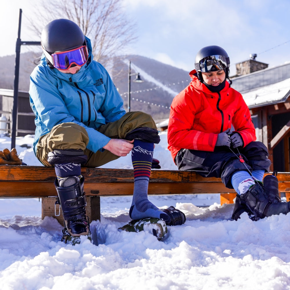 Two snowboarders seated on a bench putting on their boots and wearing Darn Tough ski socks
