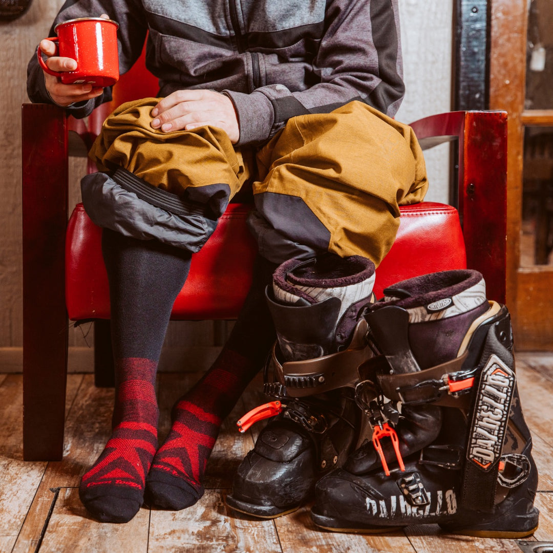 A skier in the lodge, boots off, holding a cup of hot cocoa and wearing merino wool socks for skiers