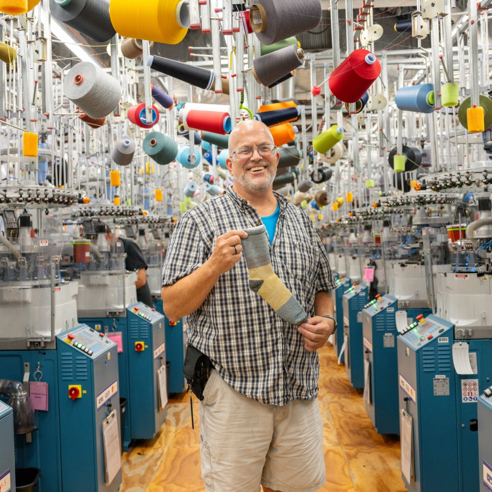 A Darn Tough employee standing at the Mill, surrounded by knitting machines, holding up his favorite sock to gift
