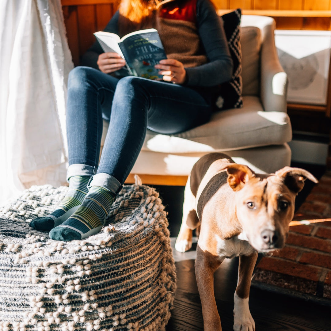 Woman reading a book wearing darn tough striped socks with her dog watching