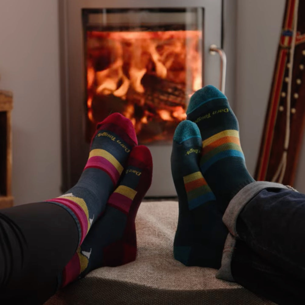 Two pairs of feet in Darn Tough socks in front of a cozy fireplace