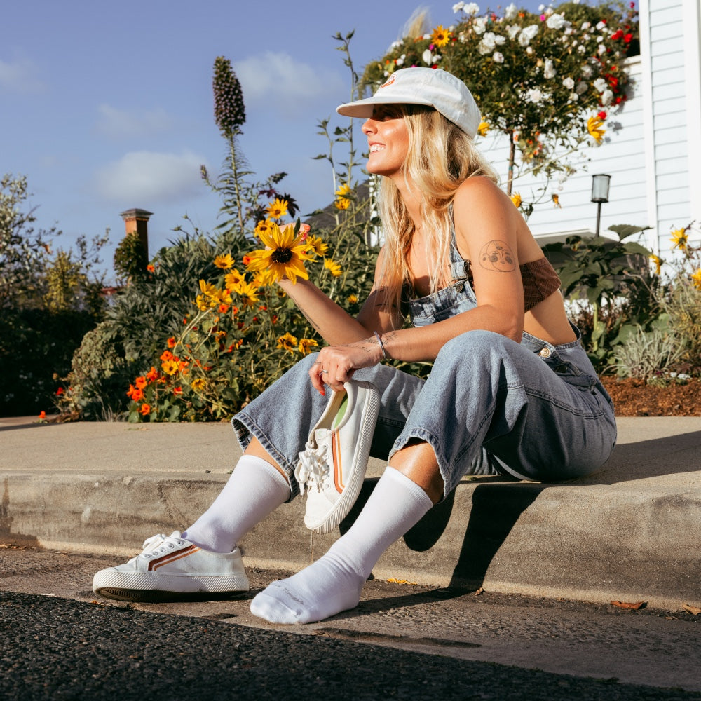 A girl wearing white crew socks, putting on low-top sneakers, and admiring sunflowers