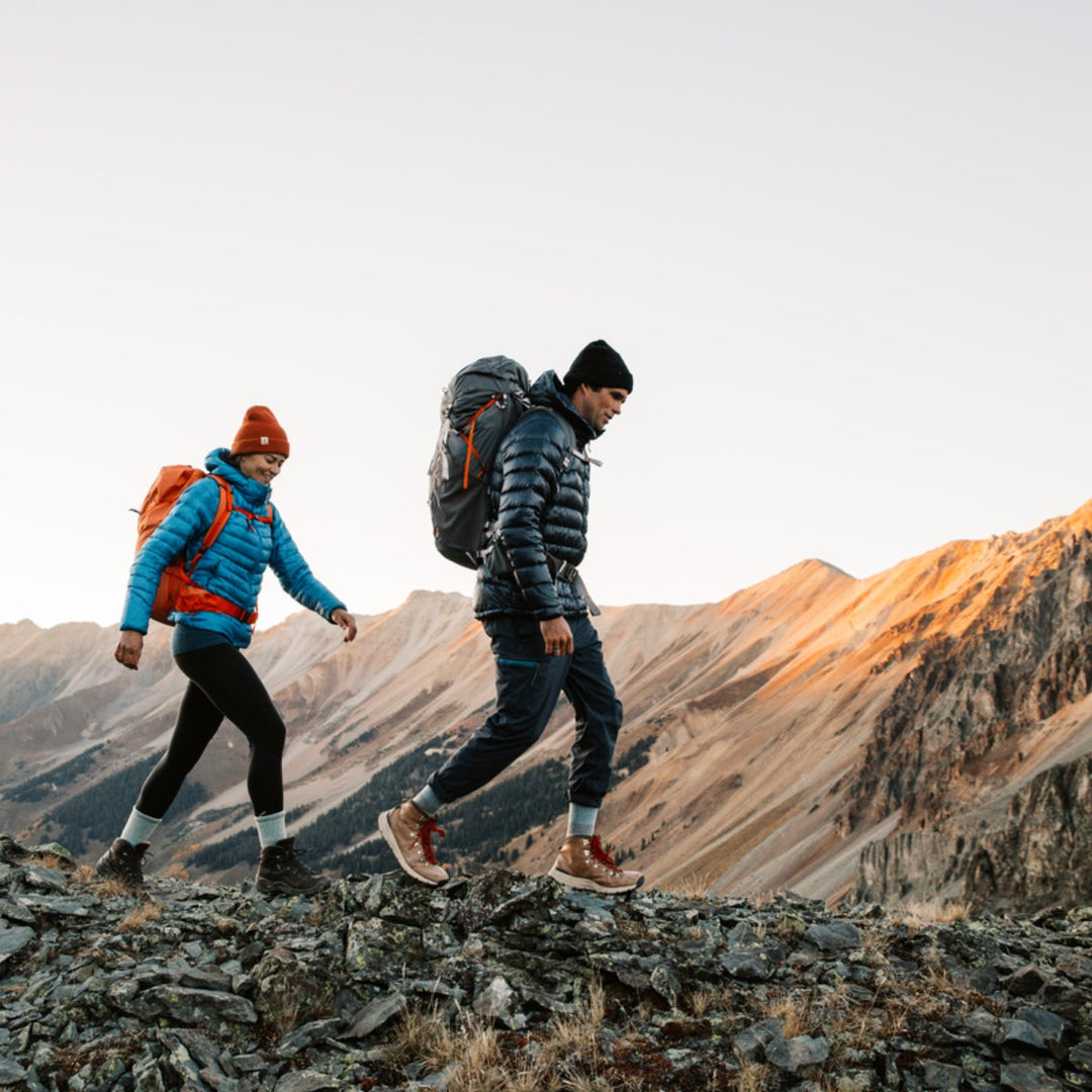 Two hikers walking across an open, rocky trail wearing puffy jackets and warm hiking socks.