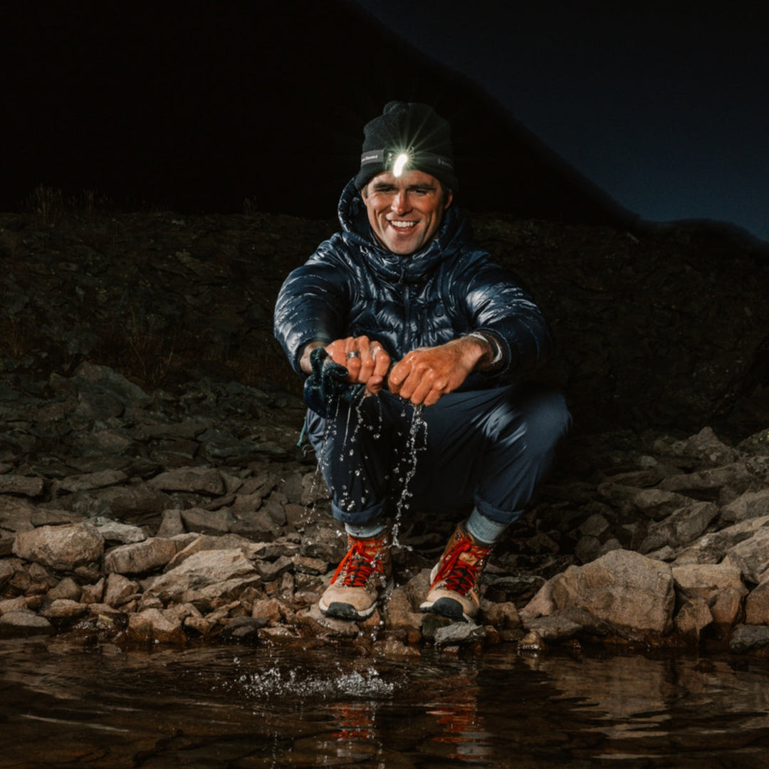 Man sitting on riverbed rocks squeezing water out of a sock over the river.