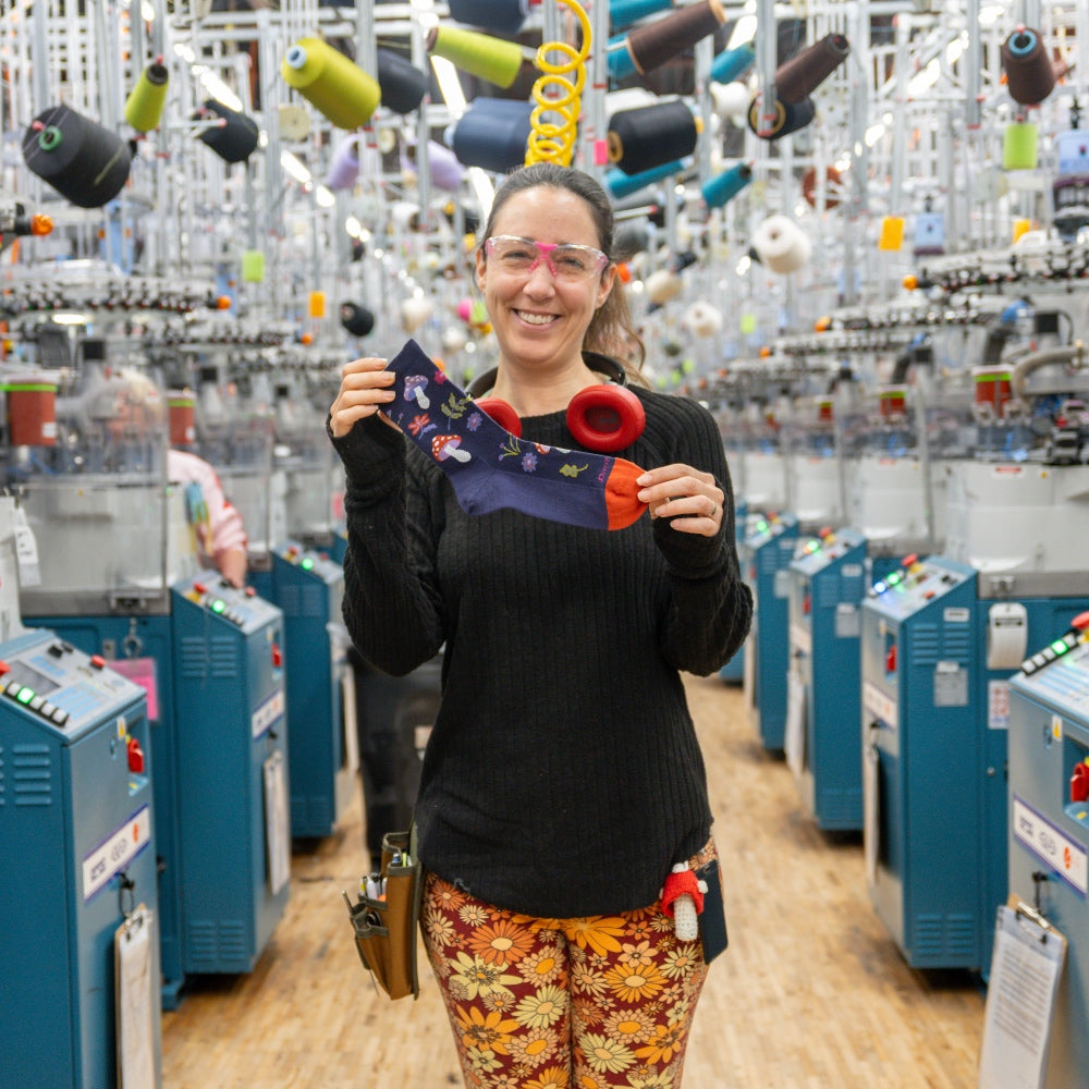 A Darn Tough employee smiling and holding up her favorite sock, surrounded by the Mill and knitting machines