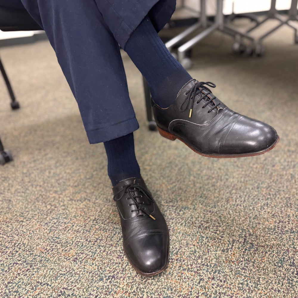 A pair of feet wearing black shoes, navy blue men's dress socks, and matching navy pants