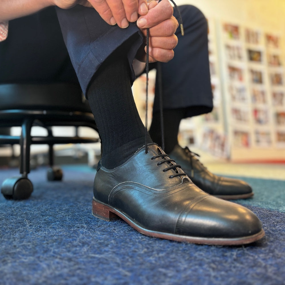 A man in suit pants putting on dress shoes over black dress socks in a crew height