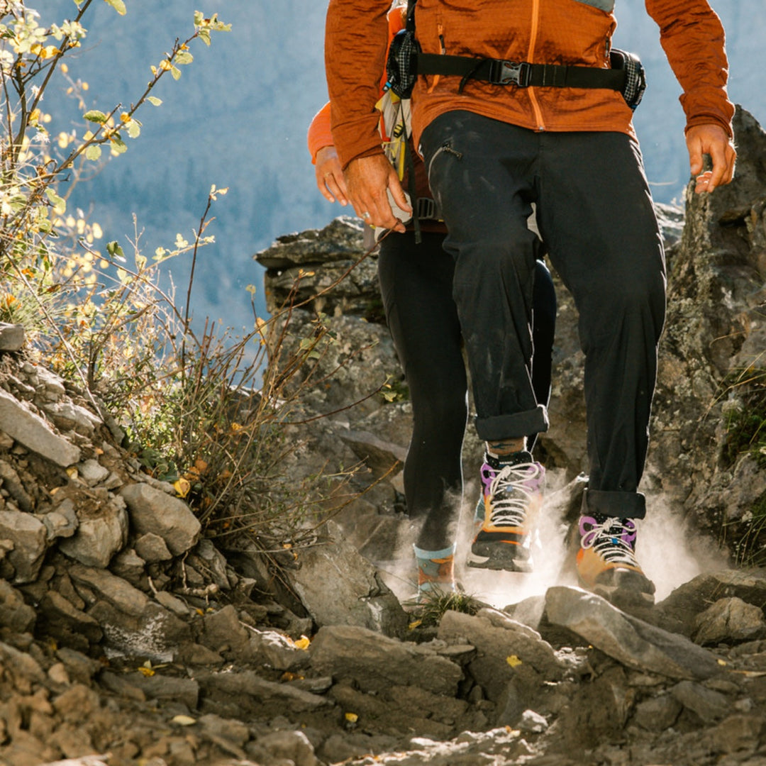 Two hikers walking across rocks on a dusty trail wearing hiking boots and hiking socks.