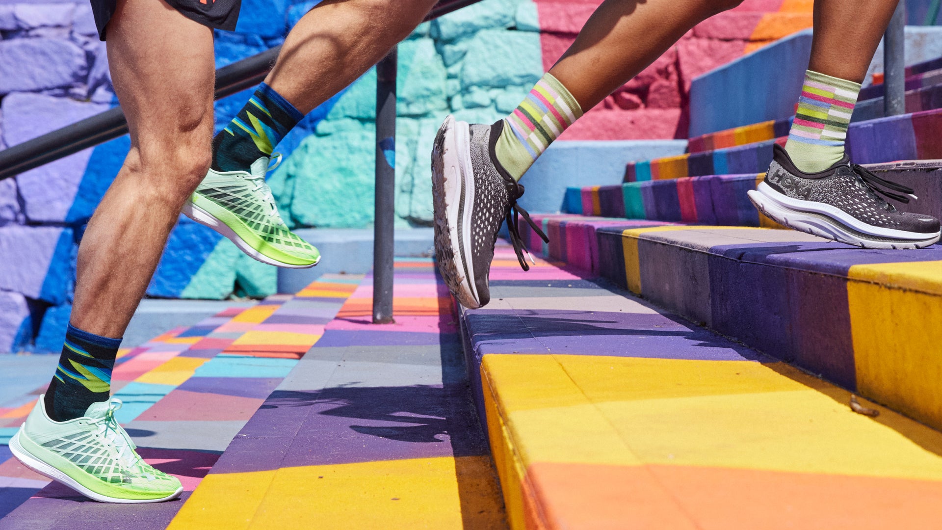 Shot of two models wearing 1056 Men's Bolt Micro Crew  Ultra-Lightweight Running Sock in black colorway and 1063 Women's Relay Micro Crew  Ultra-Lightweight Running Sock in Mint colorway running up brightly painted steps