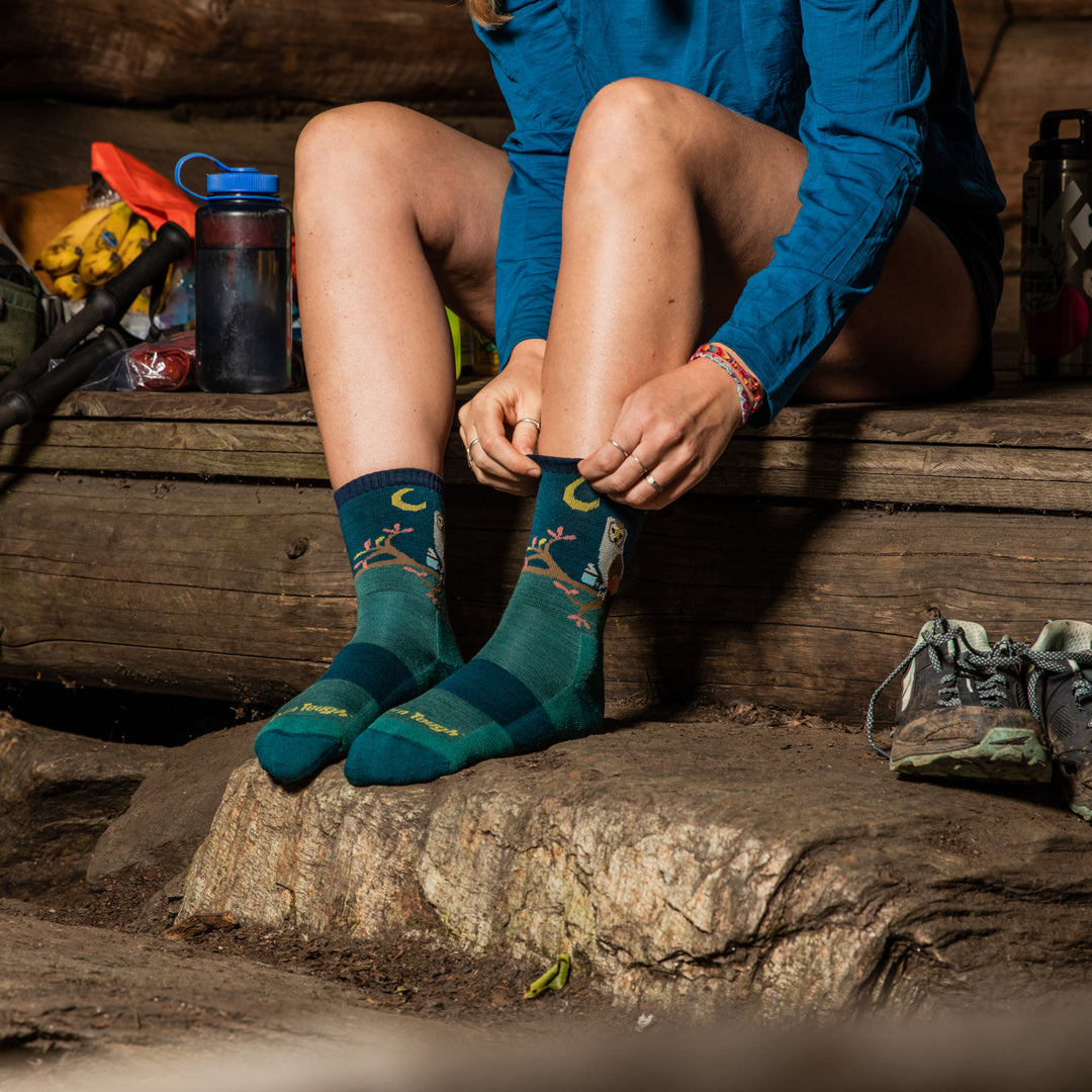 Hiker at shelter pulling on the Critter Club hiking socks in Teal with a coffee-drinking owl