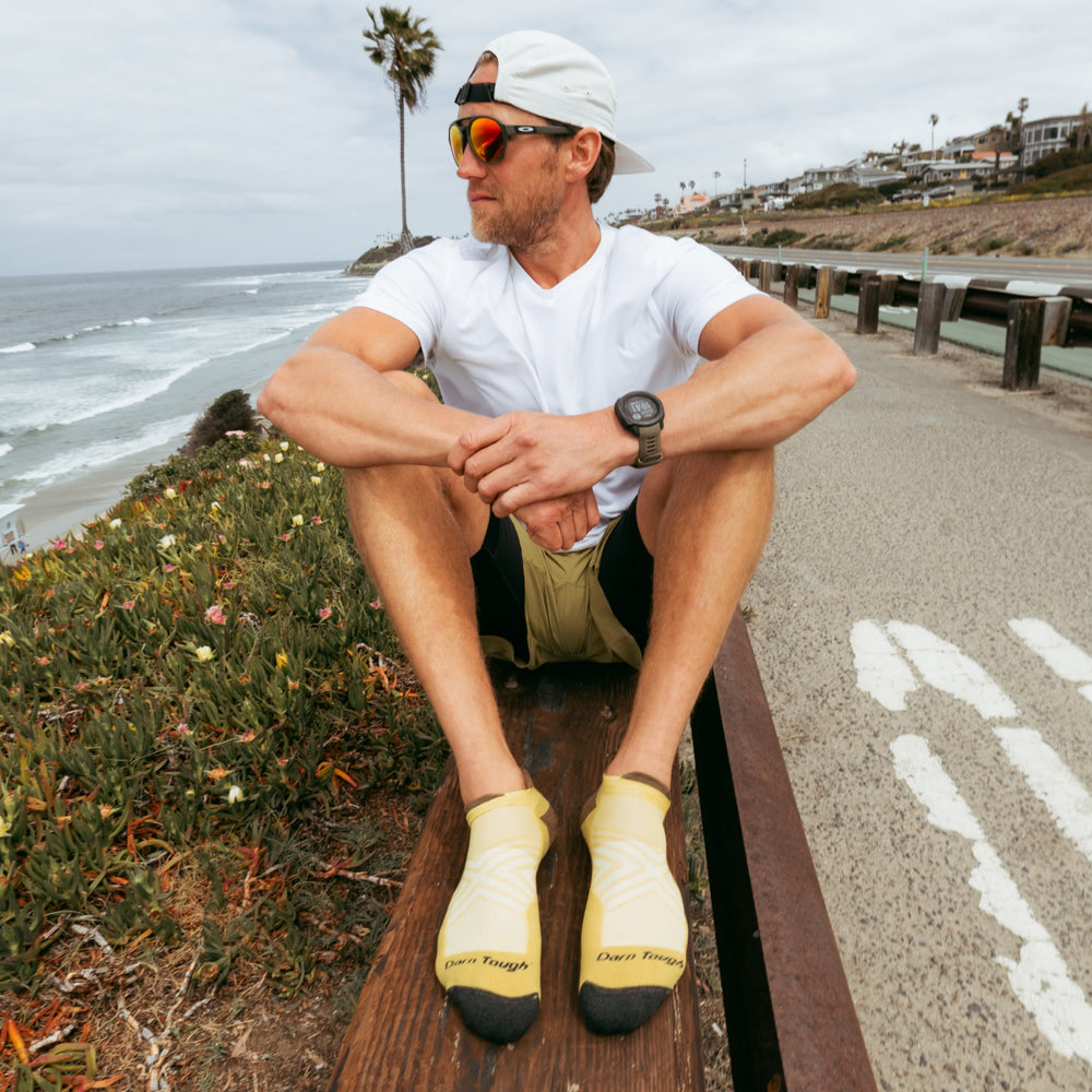Model sitting on wood bench next to ocean wearing the Men's no-show tab running sock  in sandstone colorway.