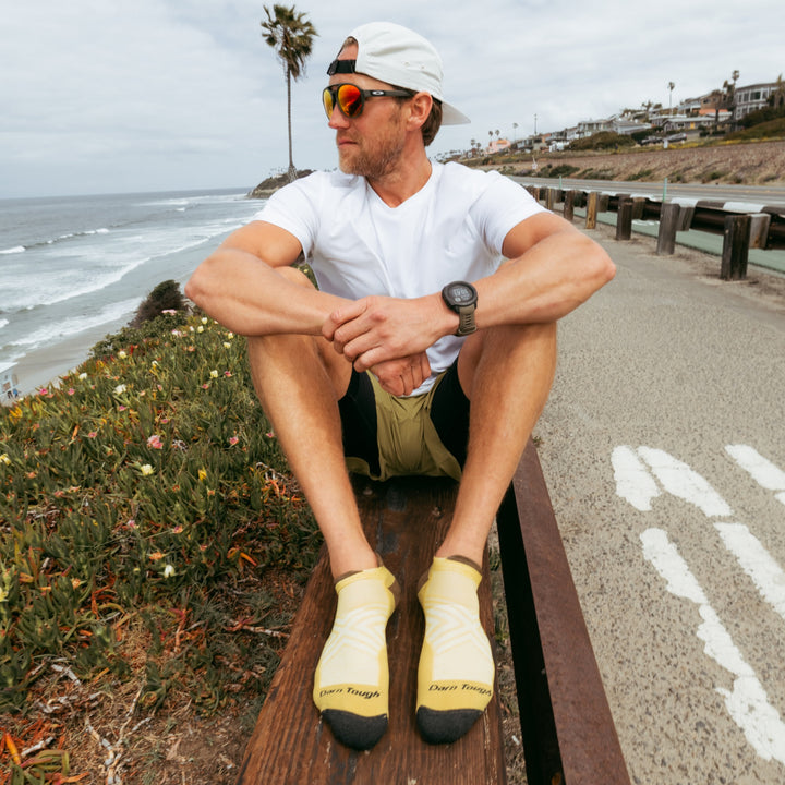 Model sitting on wood bench next to ocean wearing the Men's no-show tab running sock  in sandstone colorway.