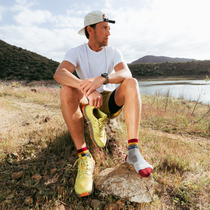 Model sitting on rock next to water holding one shoe in hand wearing the 1040 quarter running sock in skyracer Gray colorway