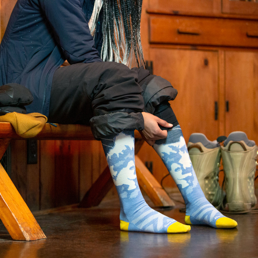 Side shot of model sitting on a bench inside putting on women's yeti over-the-calf ski sock in midnight blue