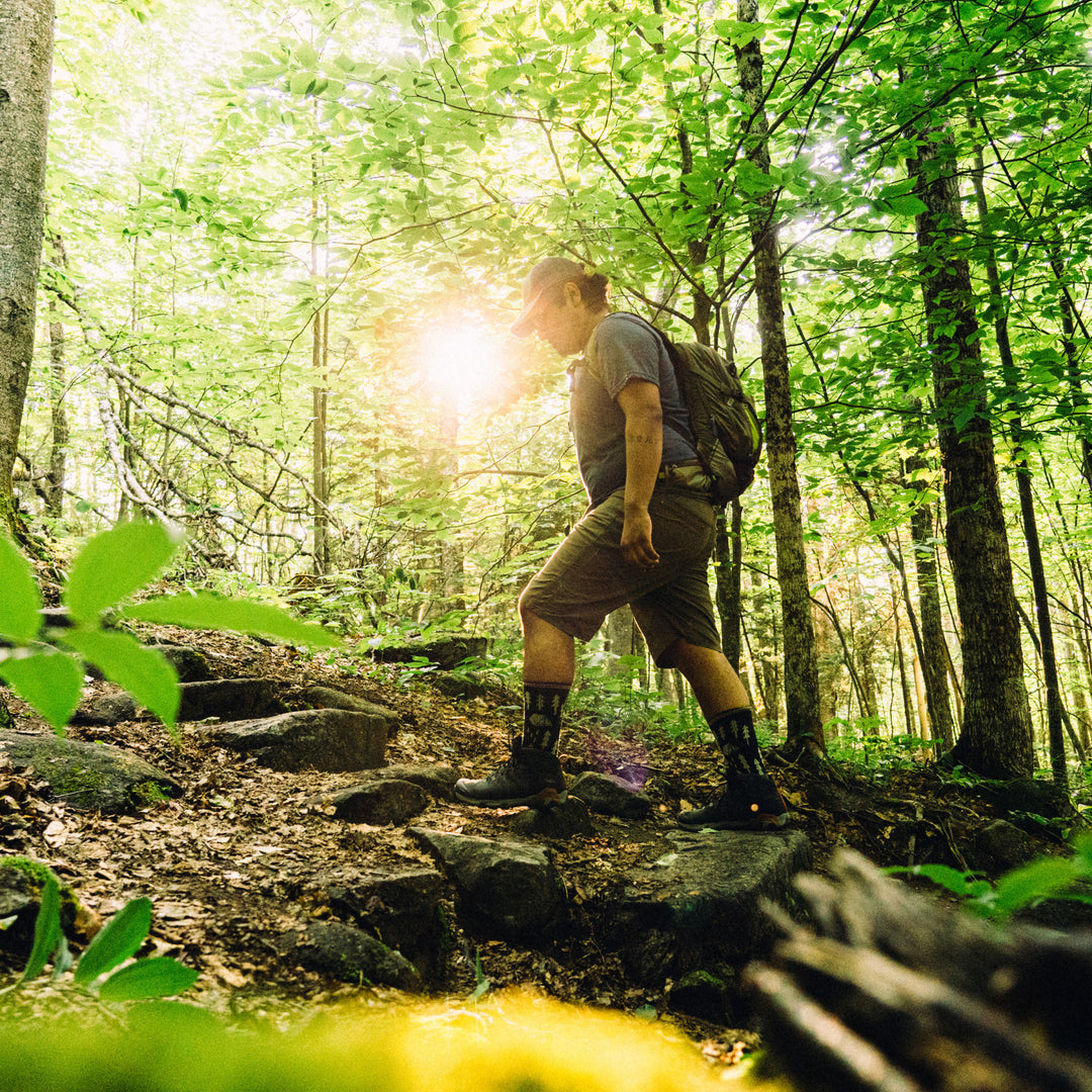 Male hiker walks through the woods wearing a backpack, hiking boots, and the ABC Boot Midweight Hiking Sock