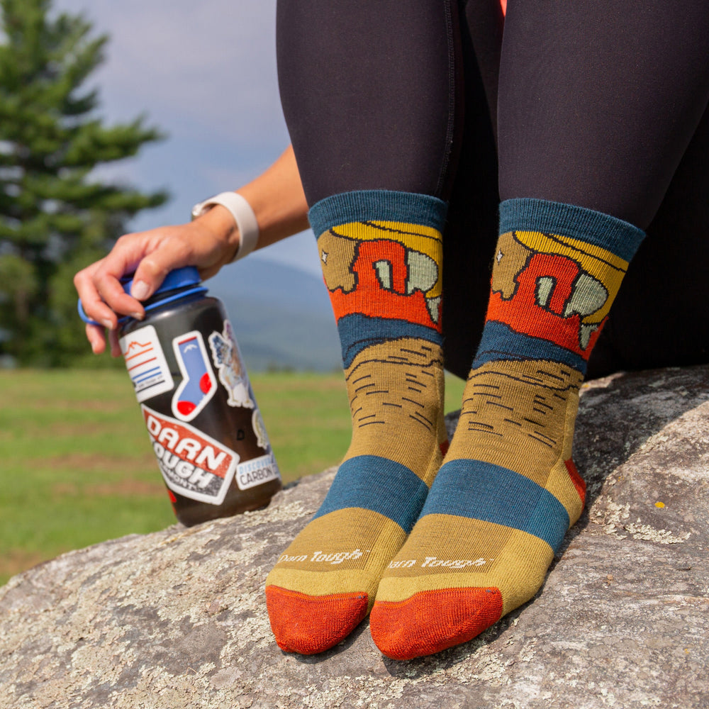 model sitting on rock with water bottle wearing the 5023 celeste micro crew in sandstone featuring yellow body orange heel/toe canyon scene on ankle