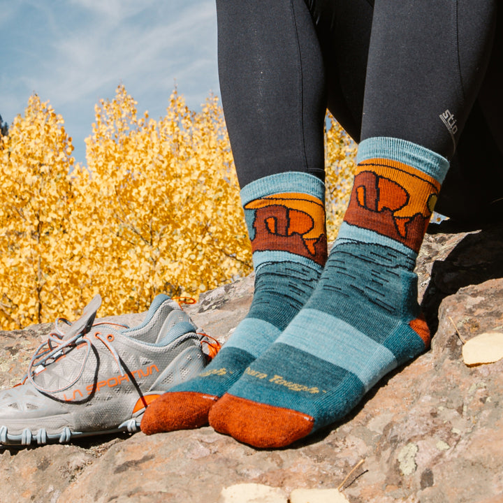 models feet on a rock wearing the 5023 celeste micro crew in teal featuring blue body and orange heel/toe with canyon scene on ankle