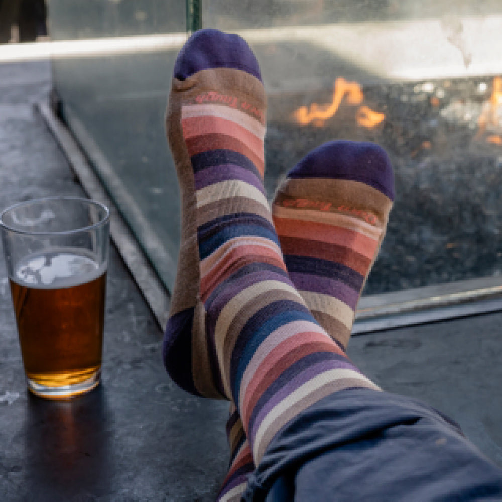 Close up shot of a model with legs crossed and wearing 6113 socks in Bark colorway with a fire pit and glass of beer in the background