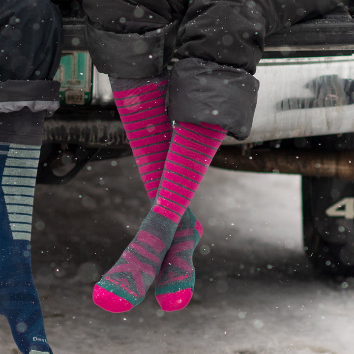 Model sitting in car trunk while it's snowing wearing women's edge over-the-calf snow sock in teal and pink