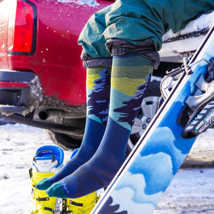 model hanging feet off back of truck with skis and boot wearing the 8014 solstice over the calf ski and snowboard sock in Eclipse