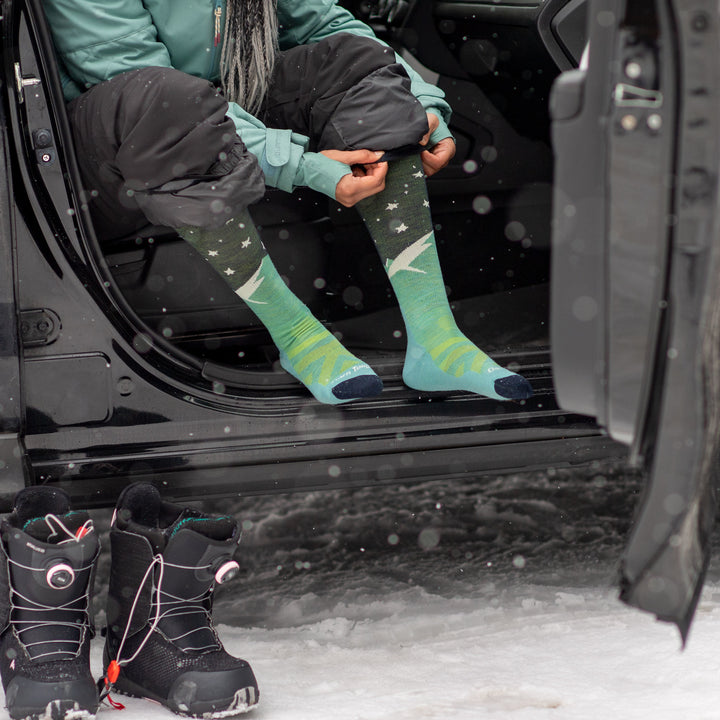 Model sitting in the backseat of a car in snow gear putting on the women's aurora over-the-calf snow sock in aqua
