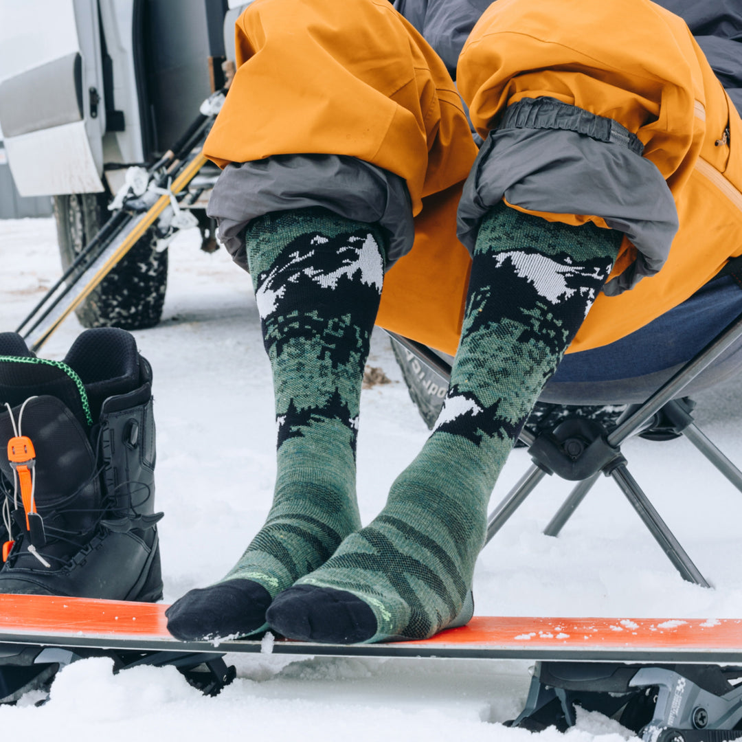 models feet resting on the bottom of snowboard wearing the 8051 cold smoke over the calf ski and snowboard sock in moss colorway
