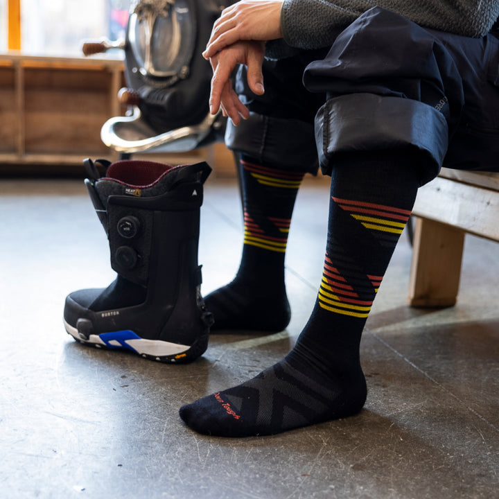 model sitting on bench with snowpant legs rolled up next to snowboard boot wearing 8061 carve OTC midweight with cushion black