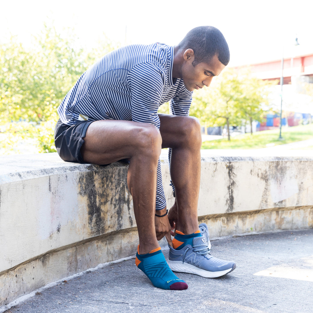 Full body shot of model sitting down putting on his shoes wearing the men's no show tab running sock in dark teal